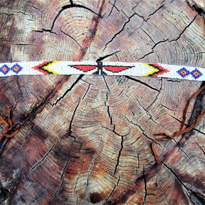 Op de afbeelding: Een wit kralen haarband met rode, gele en blauwe geometrische patronen. Het haarband heeft twee bruine leren banden.