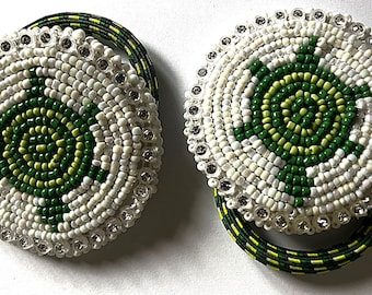 May include: Two round beaded hair ties with a white background and a green and light green circular design in the center. The hair ties are made of beads and have a clear rhinestone border.