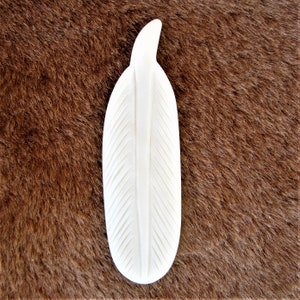 May include: White ceramic feather with a carved design. The feather is on a brown furry surface.