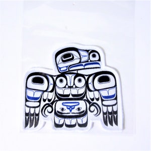 Può includere: Una toppa di tessuto bianco con un design stilizzato in nero e blu di un'aquila nativa americana. L'aquila è raffigurata in uno stile tradizionale della costa nord-occidentale con dettagli intricati e motivi geometrici.