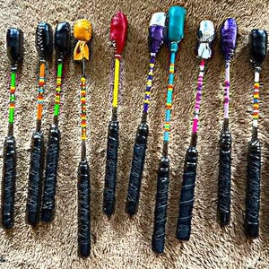 May include: A selection of handcrafted juggling clubs. Each club has a black handle with a textured grip, a shaft decorated with colourful beads, and a head in various colours and patterns, including blue, red, yellow, and purple. The clubs are arranged on a brown, textured surface.