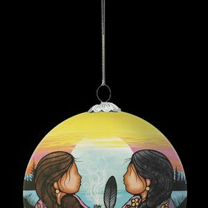 May include: A round, yellow Christmas bauble featuring a painted scene of two figures facing each other, holding a feather. The background shows a sunset over a lake and trees. The bauble hangs from a silver string.