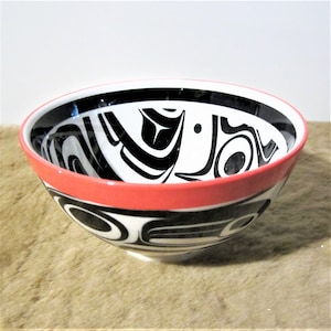 May include: A white ceramic bowl with a black and white geometric design and a red rim. The design features stylized animal figures.