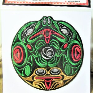 Può includere: Un cerotto rotondo ricamato in verde, rosso, oro e nero con un design stilizzato di una creatura con una testa grande e occhi. Il cerotto è su uno sfondo bianco.