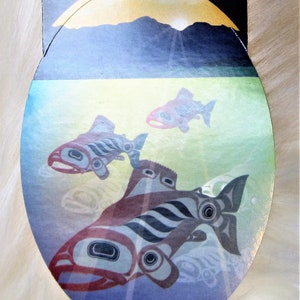 May include: A sticker featuring a stylized illustration of three salmon swimming in a blue and green underwater scene. The salmon are drawn in a traditional Tlingit style with black and red lines. The sticker is oval shaped and has a gold background with a mountain range in the distance.
