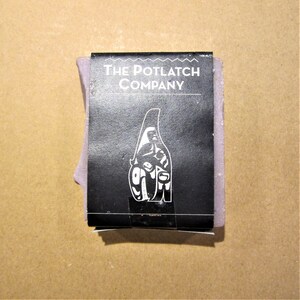 May include: A bar of lavender soap with a black label that reads "The Potlatch Company". The label features a stylised image of a person in a traditional Native American headdress.
