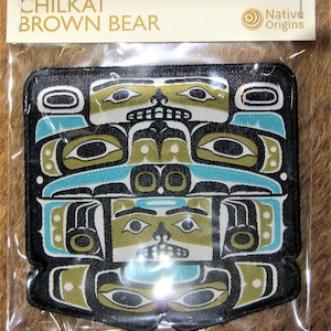May include: A rectangular patch featuring a stylized Chilkat Brown Bear design. The patch has a black border and incorporates teal, olive green, and white geometric shapes. The text "CHILKAT BROWN BEAR" is at the top.