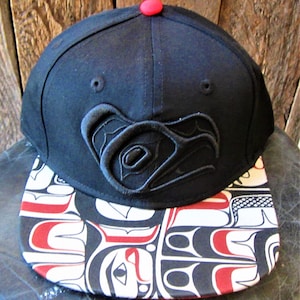 Puede incluir: Gorra de béisbol negra y roja con un diseño bordado en negro en la parte delantera y una visera estampada. La visera presenta un diseño negro, blanco y rojo.