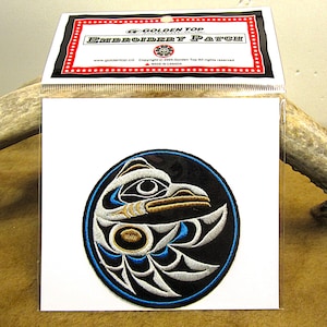 Può includere: Una toppa rotonda ricamata con uno sfondo nero e un'immagine stilizzata in bianco e oro di una testa di uccello. L'occhio dell'uccello è blu e la toppa è circondata da un bordo blu.