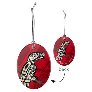 May include: Oval-shaped ornament with a red background and a stylized black and white eagle design. The ornament is suspended by a silver string. The back of the ornament is visible, with the word "back" and the text "Eagle by Corey Bulpitt, Haida".