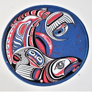 Può includere: Un cerotto rotondo blu con un designo bianco, rosso e nero di tre salmoni stilizzati che nuotano in cerchio. Lo sfondo è blu con stelle bianche.