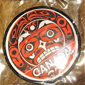 May include: A round, red, black and white embroidered patch with a stylised face and the word "CANADA" in white lettering.