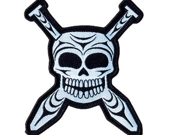 May include: A black and white embroidered patch featuring a skull with crossed knitting needles. The skull is white with black eyes and a black outline. The knitting needles are white with black details.