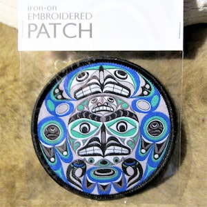 May include: Iron-on embroidered patch with a circular design featuring a stylized face with blue, green, white, and black colours. The text "iron-on embroidered patch" is printed on the white packaging.