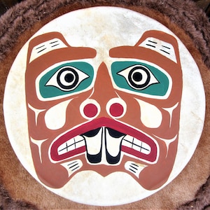 Könnte beinhalten: Eine runde, weiße Trommel mit einem bemalten Design eines Bärenkopfes. Der Bär ist in Rot, Grün, Schwarz und Weiß bemalt. Die Augen des Bären sind grün mit schwarzen Pupillen. Der Mund des Bären ist offen und hat rote Zähne. Die Nase des Bären ist rot.