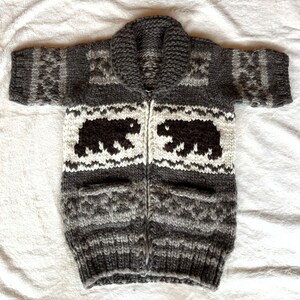 May include: A gray and white knit cardigan with a zipper closure and two pockets. The cardigan features a black bear design on the front.