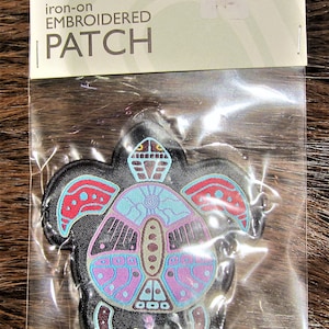 Könnte beinhalten: Aufbügel-Stickpatch mit einem farbenfrohen Schildkröten-Design. Die Schildkröte ist schwarz mit roten, blauen, pinkfarbenen und braunen Akzenten. Das Patch ist in einer durchsichtigen Plastiktüte mit einem weißen Etikett verpackt, auf dem "Aufbügel-Stickpatch" steht.