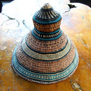 May include: Hand-woven basket hat with a conical shape. The hat features alternating bands of brown, black, and teal blue. The top of the hat is a darker shade of brown and black. The hat is made of natural fibres.