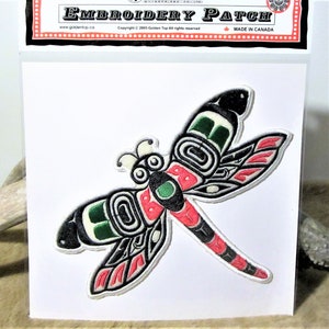 May include: An embroidered iron-on patch featuring a stylised dragonfly with black, white, red, and green colours. The dragonfly is in a Native American style with geometric patterns.