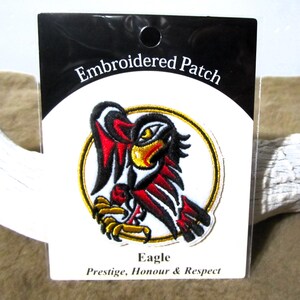 Può includere: Toppa ricamata con un designo di aquila rosso, nero e oro. La toppa è circolare e ha uno sfondo bianco. Il testo "Eagle" è sotto la toppa e il testo "Prestige, Honour & Respect" è sotto quello.