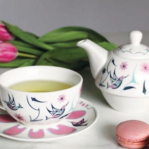 May include: A white teapot and teacup set with a floral and hummingbird design. The teapot has a lid and a spout. The teacup has a saucer. The teacup is filled with green tea. There are pink macarons in the background.