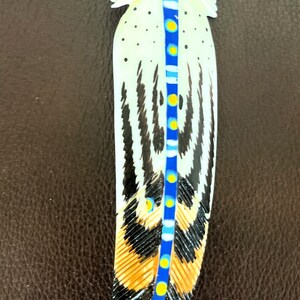 May include: A white feather with black and brown markings and a blue, white, and yellow design down the center.