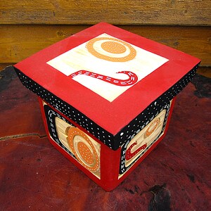 May include: A square wooden box with a red top and sides. The top has a white square with orange and beige circular designs and a red, curved design. The sides have black and white polka dot trim and beige and orange designs.