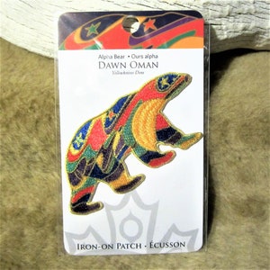 May include: Iron-on patch featuring a colourful embroidered bear with stars and the text "Alpha Bear - Ours alpha DAWN OMAN Yellowknives Dene" and "IRON-ON PATCH - ÉCUSSON".
