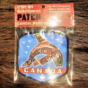 May include: Iron-on embroidered patch featuring a colourful stylised orca whale design with the word "CANADA" on a blue background. The patch is packaged in a clear plastic bag with a header card that reads "Iron on Embroidered Patch Canadian Memories Art Work Designed by Destiny Hoostie from White Bear First Nation 29".