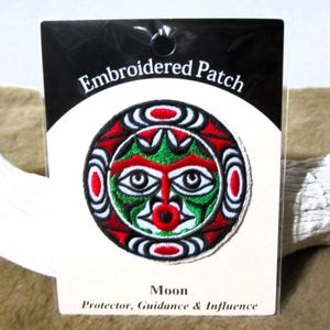 Può includere: Toppa ricamata con un designo stilizzato di volto lunare in rosso, verde, bianco e nero. Il testo "Embroidered Patch" è in cima alla carta e "Moon Protector, Guidance & Influence" è in fondo.