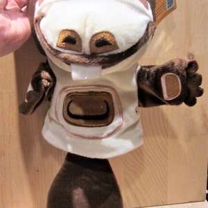 May include: Brown and white plush puppet with a large toothy grin. The puppet has a brown nose, brown eyes, and a white mouth with a single tooth. The puppet is wearing a white shirt with a brown pocket and brown sleeves. The puppet has a brown tail.