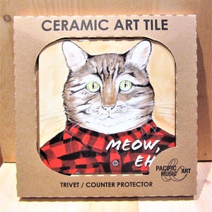 Può includere: Un sottopentola in ceramica con un'illustrazione di un gatto di cartone che indossa una camicia a quadri rossi e neri. Il testo "MEOW, EH" è stampato sulla piastrella. La piastrella è confezionata in una scatola di cartone con le parole "CERAMIC ART TILE" e "TRIVET / COUNTER PROTECTOR" stampate su di essa.