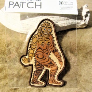 Könnte beinhalten: Gestickter Aufnäher mit einem stilisierten Bigfoot-Design in Braun- und Beigetönen. Der Aufnäher hat ein detailliertes, tribal inspiriertes Muster und blaue Augen. Der Aufnäher befindet sich in einer transparenten Plastikhülle mit einem weißen Etikett "PATCH".