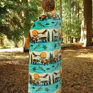 May include: A beige and turquoise blanket with a repeating pattern of wildlife, including bears, moose, eagles, and fish. The blanket has a nature-inspired design with a sun and tree motif.