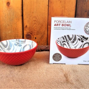 May include: A red porcelain bowl with a textured exterior and a white interior featuring a black and gray Indigenous design. The bowl is 6 inches in diameter and holds 16 ounces. The bowl is packaged in a white box with the words "Porcelain Art Bowl" printed on it.