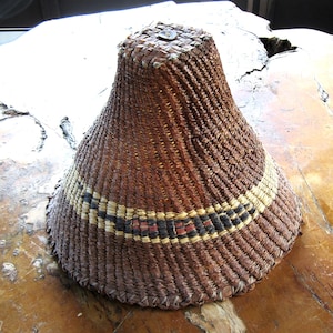May include: A woven brown hat with a flared shape. The hat features a band of black, red, and yellow geometric patterns. The hat is made of natural fibres and has a square top with a small metal detail.