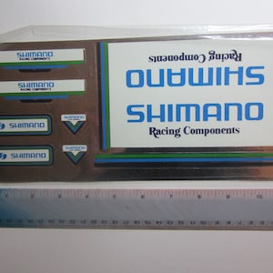 Könnte beinhalten: Eine Blatt mit Vintage-Aufklebern für Shimano-Rennradkomponenten. Die Aufkleber sind in verschiedenen Größen und zeigen das Shimano-Logo in Blau und Weiß mit dem Text "Racing Components".