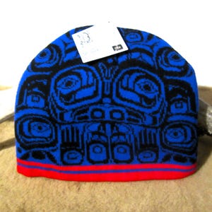 May include: Blue knit hat with a black and blue patterned design featuring stylized faces. The hat has a red brim.