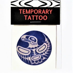 Puede incluir: Un tatuaje temporal que presenta un diseño estilizado de un pájaro con las alas extendidas en azul y blanco. El tatuaje está empaquetado en una manga de plástico transparente con una etiqueta negra y roja que dice "TATUAJE TEMPORAL".