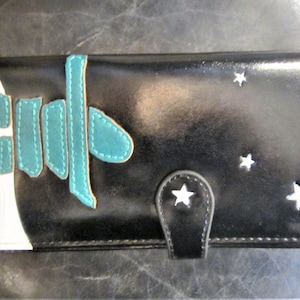 Op de afbeelding: Zwarte portemonnee met witte en turquoise accenten. De portemonnee heeft een wit gedeelte met turquoise stiksels en een turquoise ontwerp. Witte steruitsparingen en een stervormige sluiting voegen details toe.