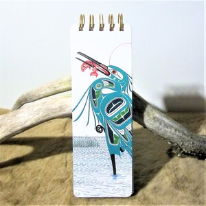 May include: A small, white spiral bound notebook featuring a colourful illustration of a blue and green bird with a red fish in its beak. The bird is standing in blue water with green reeds in the background.