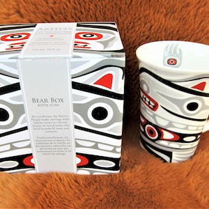 May include: A white ceramic mug with a black, red, and gray bear design. The mug is sitting on a brown furry surface next to a box with the same design. The box has the text "BEAR BOX BOITE OURS" on the front. As a tradition, the Native People make carvings with family crests or cultural stories on wood boxes that hold household items and traditional treasures. Traditionally, the Autochtones adopt the emblems of the family on the boxes that hold the containers for the food and treasures of the marriage.
