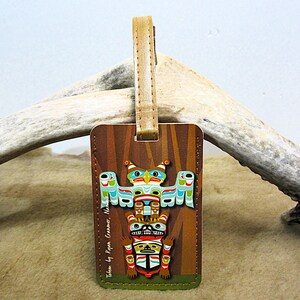 May include: A rectangular luggage tag with a vibrant totem pole design, including an owl, bear, and human figures. The tag has a brown background with vertical lines and a tan leather strap. The text "Totem by Ryan Cranmer, Native" is printed on the tag.