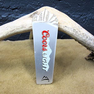 May include: A silver Coors Light beer tap handle with a mountain design. The handle features the Coors Light logo in red and white, and a mountain graphic. The top of the handle is carved with a mountain range.