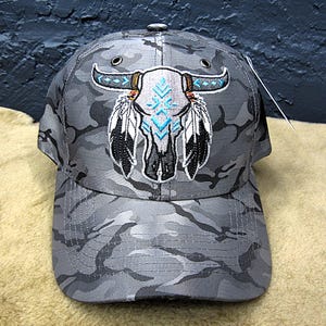 May include: Gray camouflage baseball cap featuring an embroidered design of a bull skull with turquoise accents and feather details. The cap has a curved brim and a button on top.