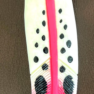 May include: A decorative feather-shaped object with a vibrant design. The feather features a bright pink central stripe, black polka dots, and black accents. The base color is a creamy white, and the feather is set against a brown background.
