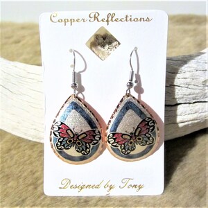 May include: Copper teardrop earrings with a butterfly design. The earrings are silver with blue and gold accents. The butterfly has red, white, and blue wings. The earrings are designed by Tony.