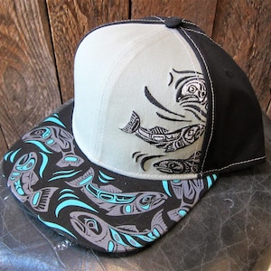 Op de afbeelding: Een zwarte en grijze baseballpet met een grijze kroon en een zwarte klep. De klep heeft een kleurrijk ontwerp van gestileerde zalm in tinten blauw, grijs en zwart.