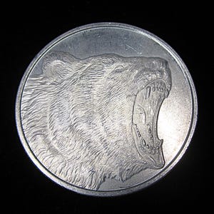 May include: A silver coin with a detailed relief of a bear's head, its mouth open to reveal teeth. The coin is circular and has a textured surface, with the bear's fur intricately depicted. The coin's design is striking.