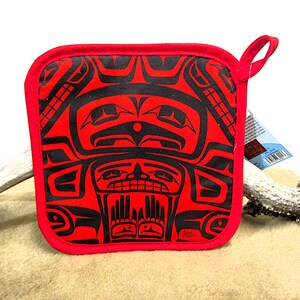 May include: Red square pot holder with a black and white design of a Native American art pattern. The design features a face with eyes, nose, and mouth. The pot holder has a loop handle.